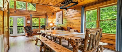 A rustic cabin interior with a large wooden dining table, benches, and a cozy seating area.