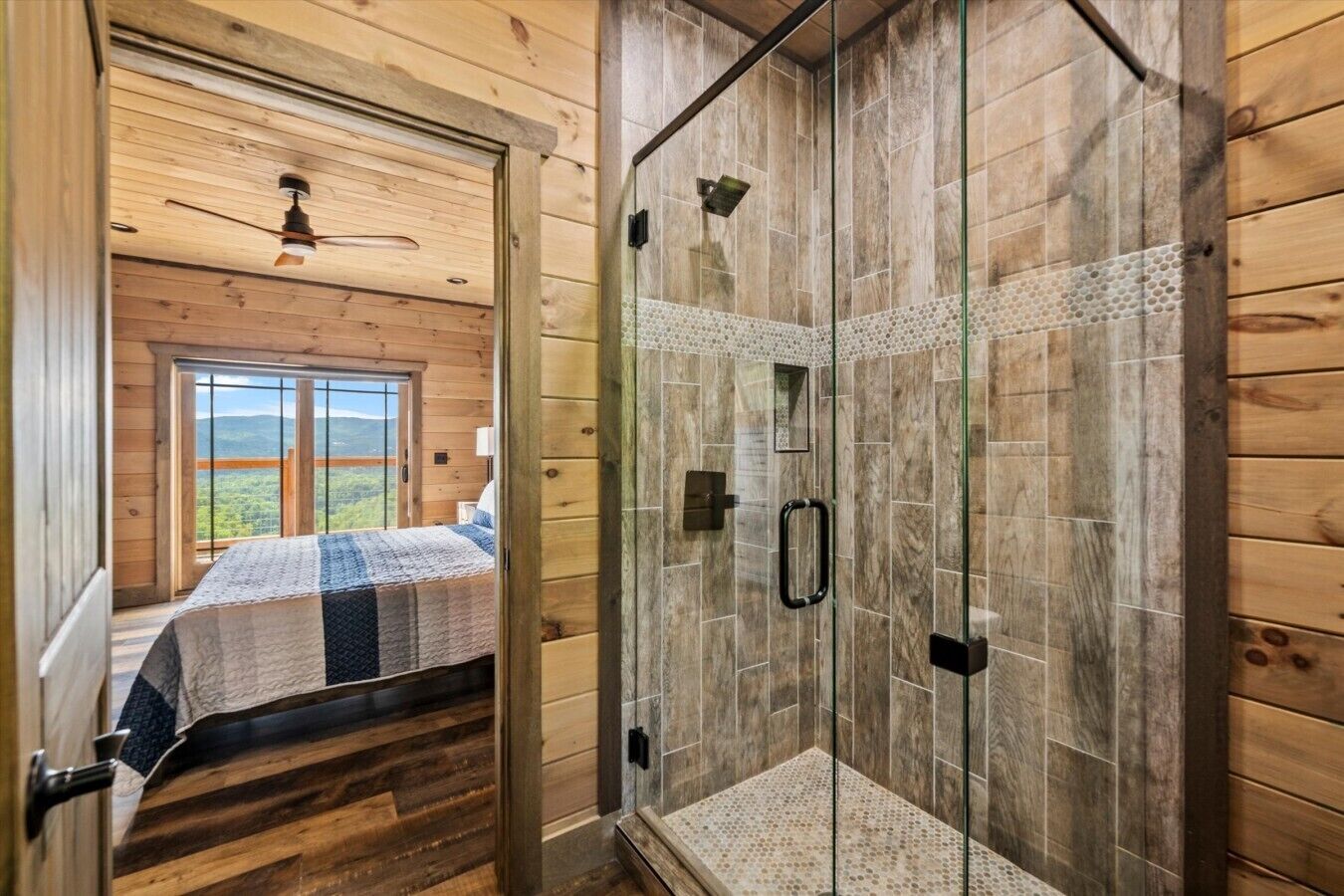 A wooden cabin interior with a glass-enclosed shower, a bed with a view, and a wooden ceiling.