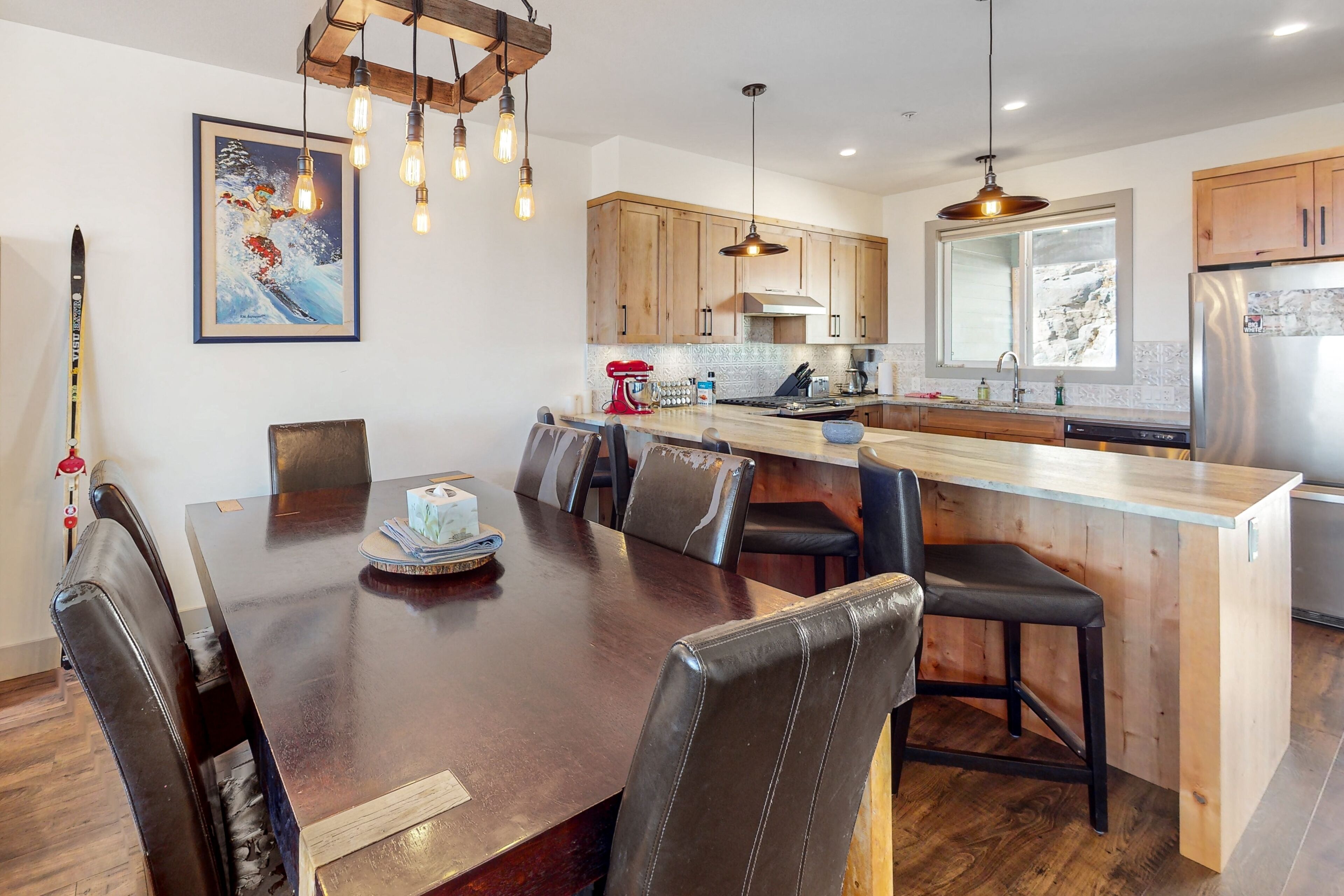 A modern kitchen with wooden cabinets, a stainless steel refrigerator, and a dining area with a wooden table and leather chairs.
