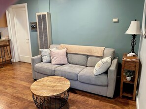 A living room with a grey sofa, a round wooden coffee table, a small wooden side table with a lamp, and a wall-mounted air conditioning unit. - Soothing 1-bedroom house in charming Kings Beach perfect for getaway (Kings Beach)