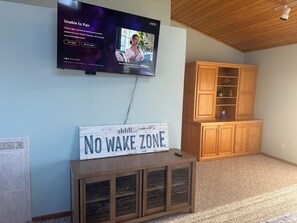 A living room with a television mounted on the wall, a wooden cabinet, and a sign on a console table that reads 'NO WAKE ZONE'.