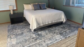Quarto com cama, mesa de cabeceira, abajur, janela com persianas e tapete.