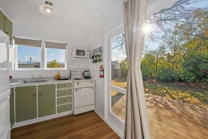 A kitchen with a microwave, stove, and cabinets, a window with a view of greenery, and a curtain. - Tui Shack (Kinloch)