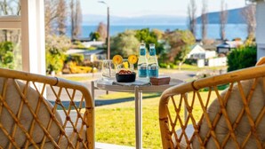 A balcony with wicker chairs, a small table, and a view of the sea and houses. - Tui Shack (Kinloch)