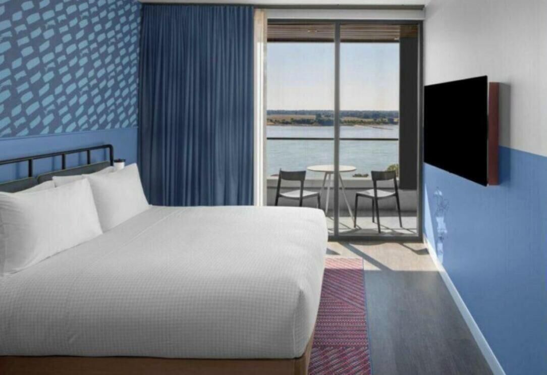 A hotel room with a bed, a TV, a balcony with a view of the water, and a patterned wall.