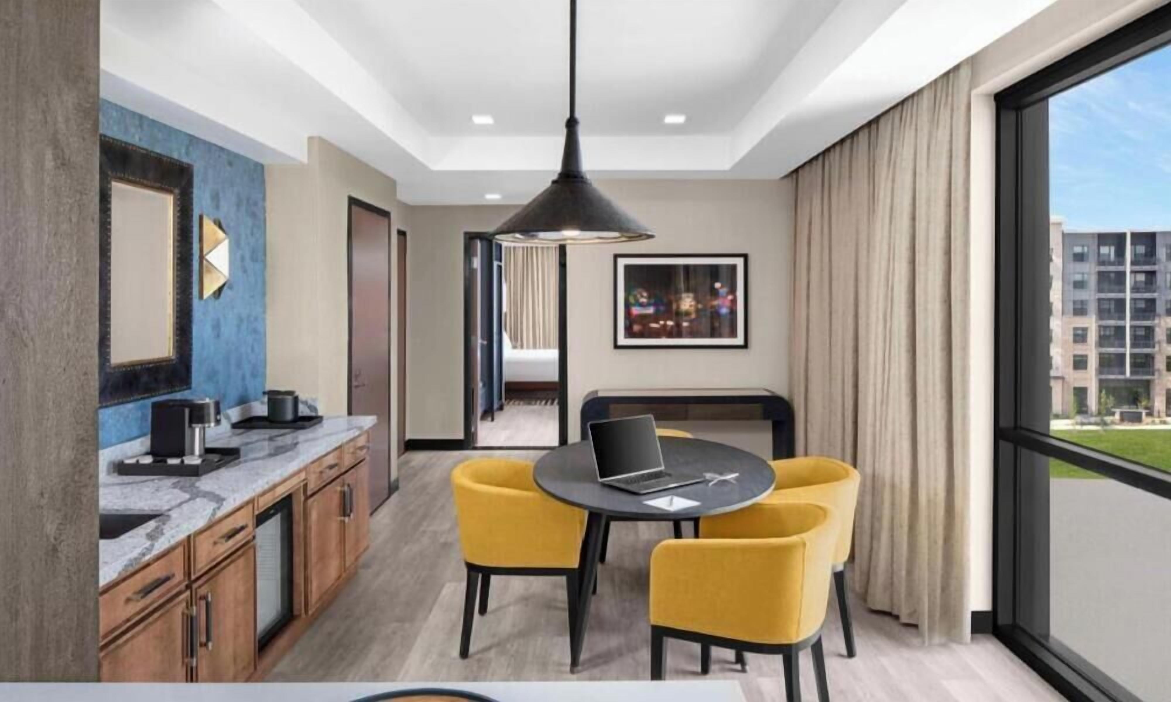 A modern hotel room with a kitchen area, a dining table with yellow chairs, a large window with a view of buildings, and a ceiling light fixture.