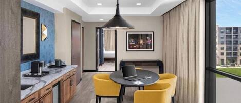 A modern hotel room with a kitchen area, a dining table with yellow chairs, a large window with a view of buildings, and a ceiling light fixture.