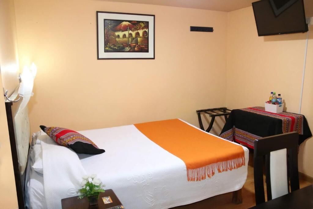Una cama individual con colcha blanca y naranja, una mesita de noche con un florero, y un cuadro en la pared.
