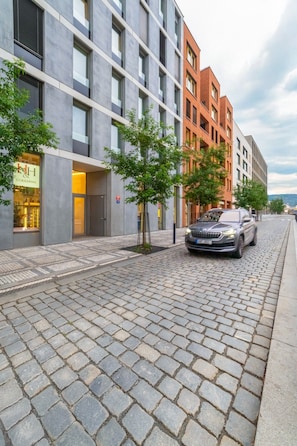 A modern building with a mix of grey and red brick facades, a paved cobblestone street, and a parked car. - The Elegant White Suite in SmichovCity Residence (Prague)
