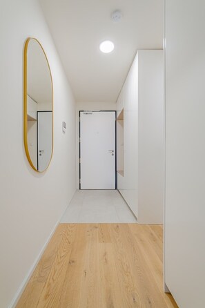 A hallway with a wooden floor, a white door at the end, and a large mirror on the left wall. - The Elegant White Suite in SmichovCity Residence (Prague)