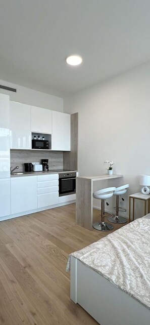 A modern kitchen with white cabinets, a microwave, and a coffee maker. There is a small bar area with stools and a table. - The Elegant White Suite in SmichovCity Residence (Prague)