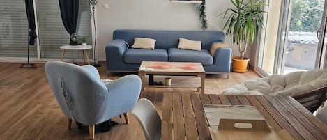 Een moderne woonkamer met een blauwe bank, houten vloeren en een groot raam dat natuurlijk licht binnenlaat.
