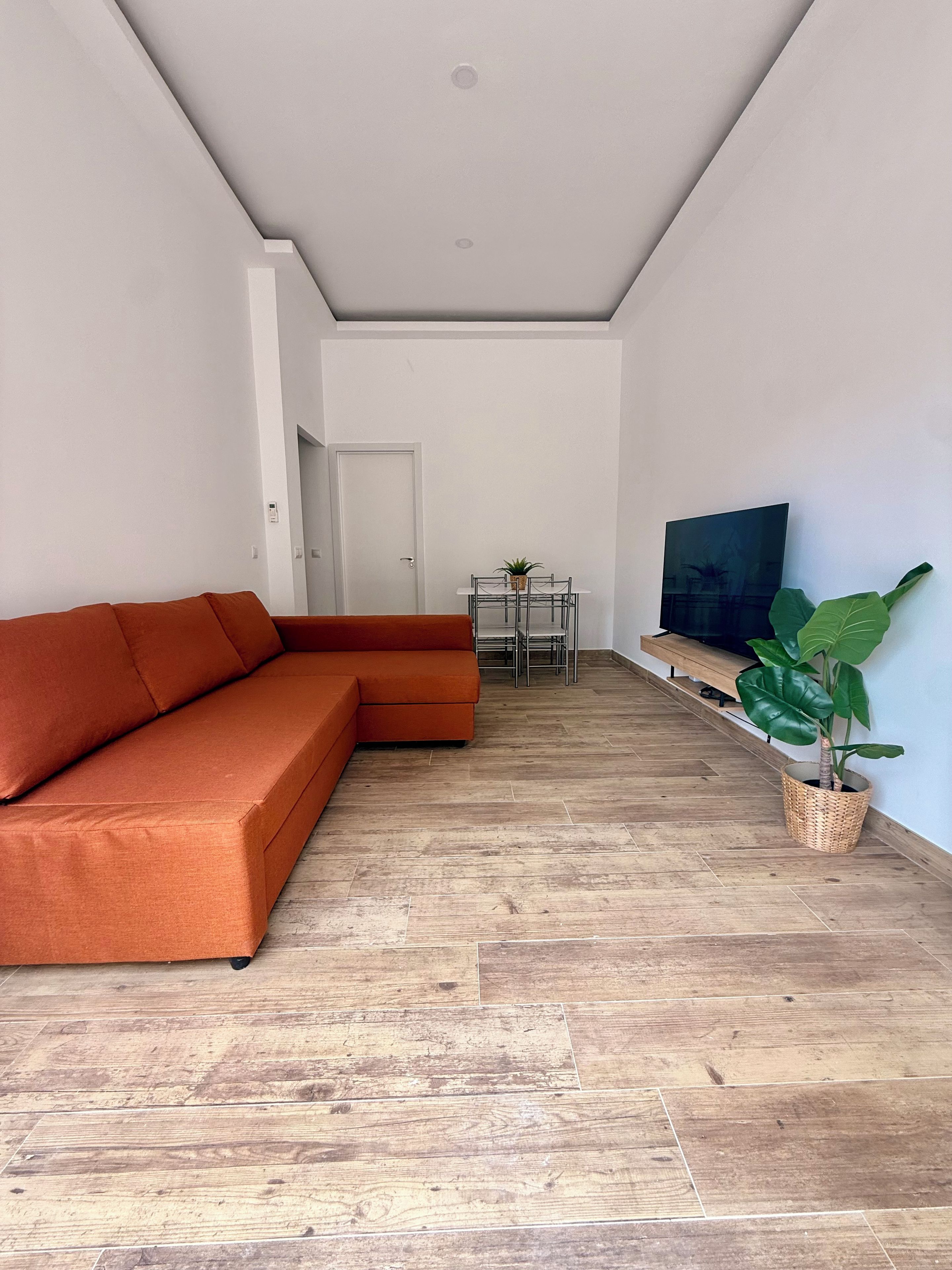 A living room with a sectional sofa, a dining table with chairs, a television, and a potted plant.