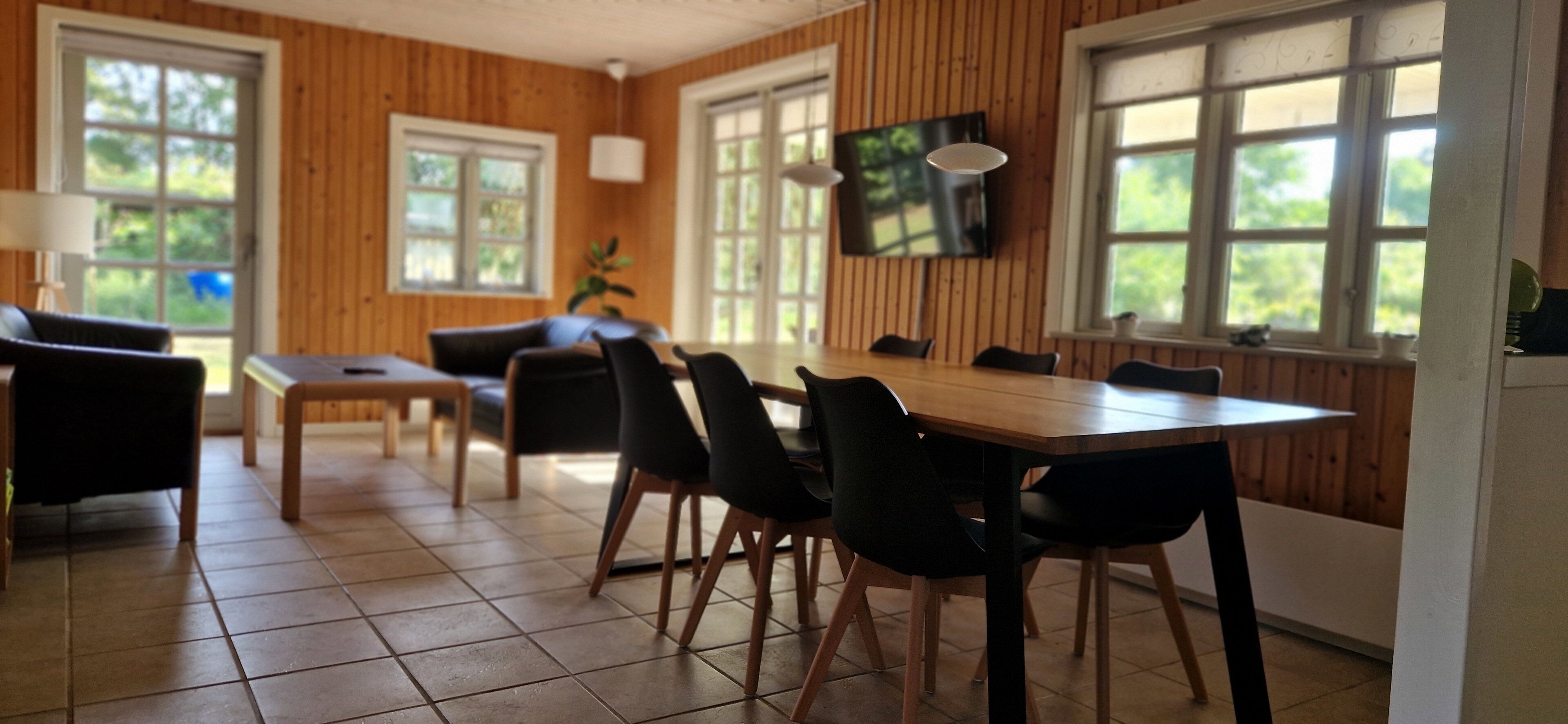 A dining area with a wooden table and chairs, a television mounted on the wall, and large windows with a view of greenery.