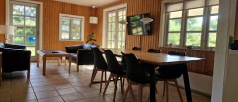 A dining area with a wooden table and chairs, a television mounted on the wall, and large windows with a view of greenery.