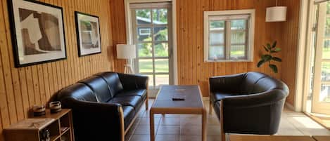 A room with a wooden paneled wall, a black leather sofa, a wooden coffee table, and a single chair. There are two framed pictures on the wall, a lamp, and a window with a view of greenery outside.