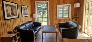 Ein Zimmer mit einer holzverkleideten Wand, einem schwarzen Ledersofa, einem Holztisch und einem einzelnen Sessel. An den Wänden hängen zwei gerahmte Bilder, eine Lampe und ein Fenster mit Blick auf grüne Natur.