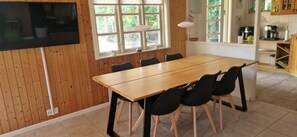 A dining area with a wooden table and black chairs, a TV mounted on the wall, and windows with a view of greenery.