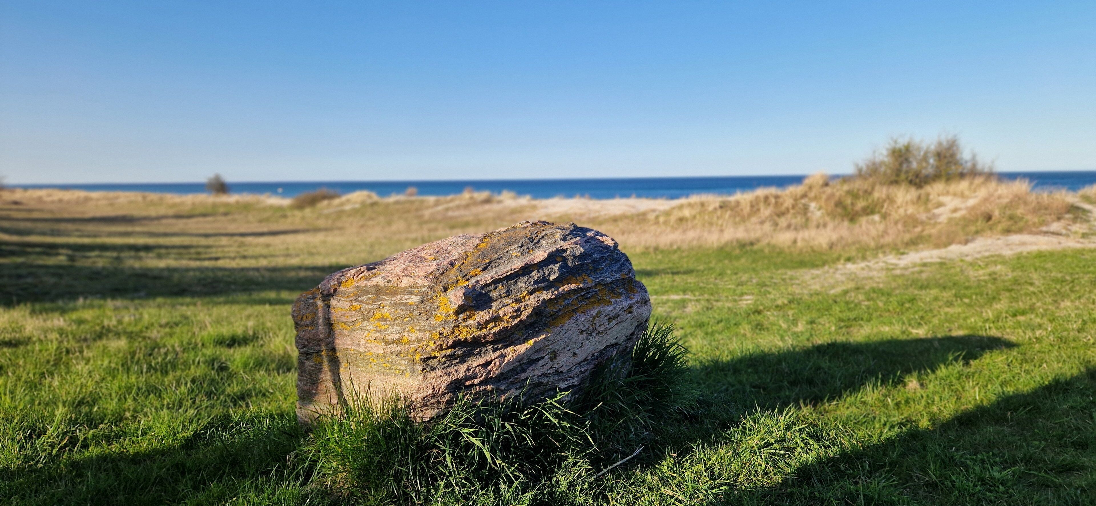 Ein großer Felsen in einer grünen Wiese, im Hintergrund ein klarer Himmel und das Meer.