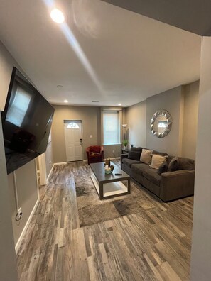 A living room with a flat-screen TV mounted on the wall, a gray sofa, a coffee table, and a wooden floor. - Experience the Charm of Hyde Park in this Renovated St. Louis Retreat (St. Louis)