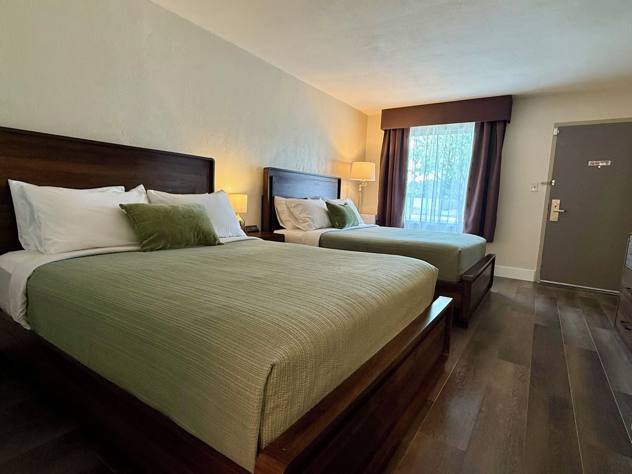 Une chambre d’hôtel avec deux lits, une tête de lit en bois, une table de chevet, une lampe, une fenêtre avec des rideaux et une porte.