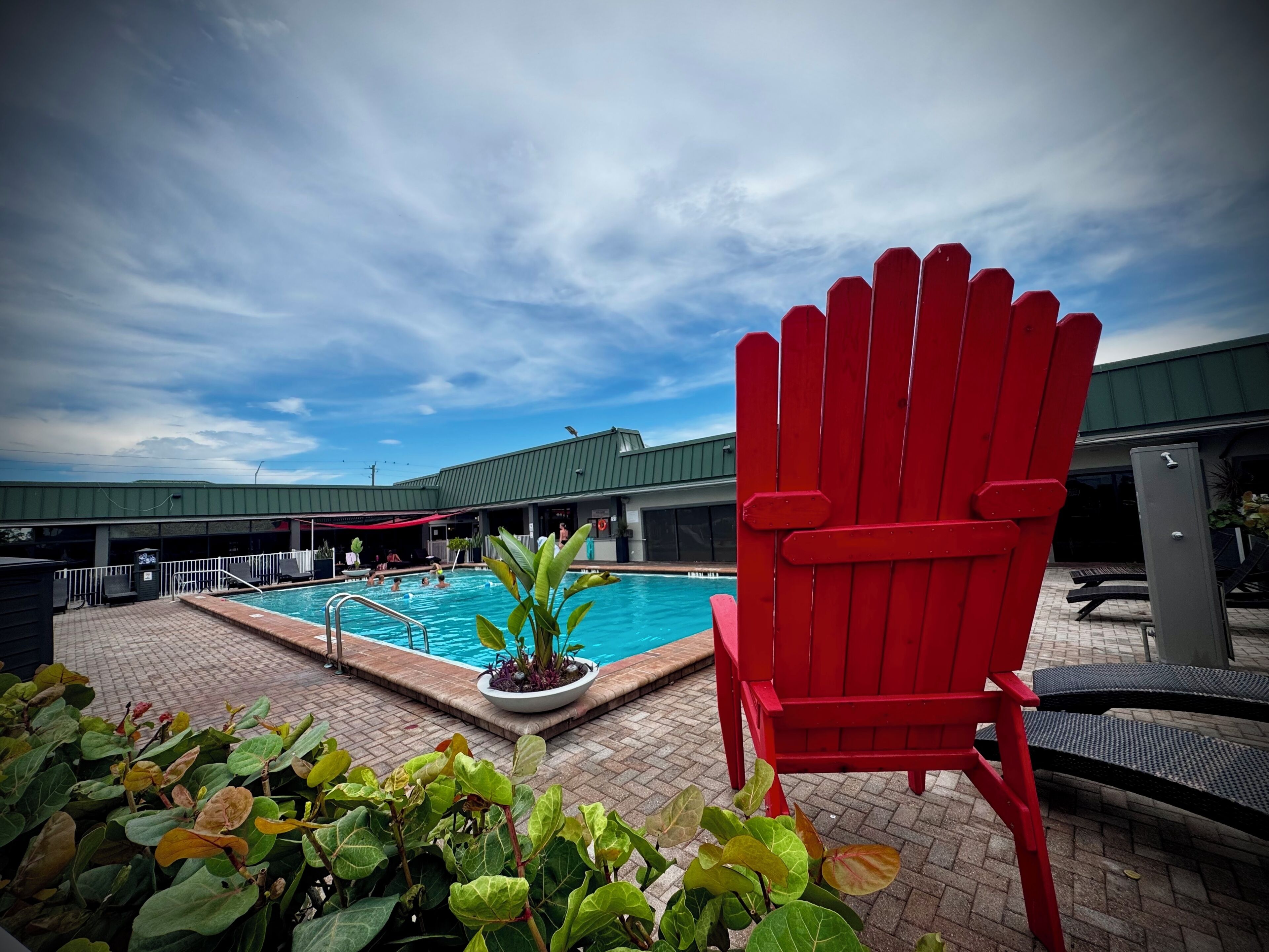 A swimming pool with a red chair in the foreground, surrounded by potted plants and a building with a green roof in the background.