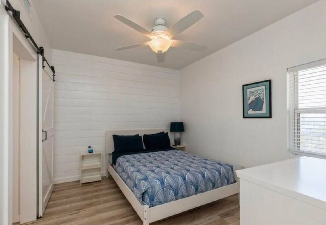 Une chambre à coucher comprenant un lit, un ventilateur de plafond, une table de chevet et une commode, une fenêtre et une porte.