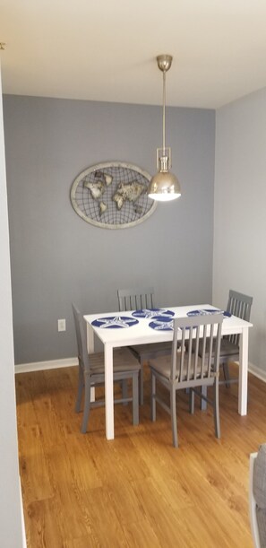 Een eetruimte met een witte tafel, grijze stoelen, een hanglamp en een decoratief object aan de muur.