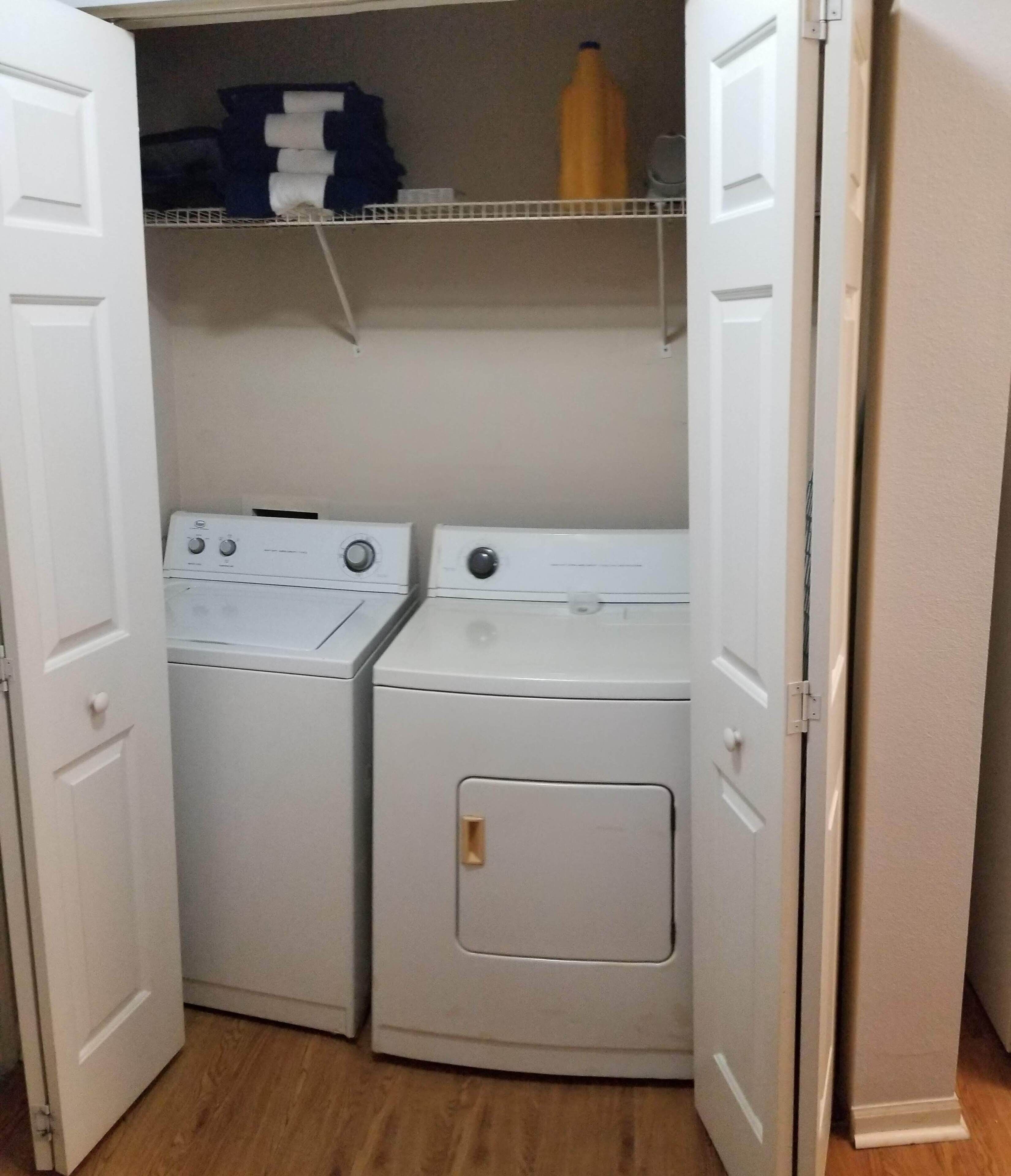 A laundry room with two white front-loading washing machines and a dryer, with a shelf above containing folded clothes and a bottle.