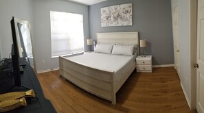 Ein Schlafzimmer mit einem Bett, Nachttischen, einem Schreibtisch mit Computer, einem Fernseher und einem Fenster mit Jalousien.