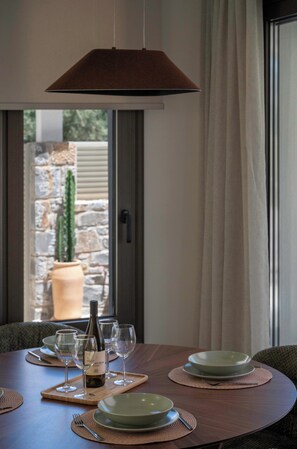 A dining table with plates, wine glasses, and a bottle, set against a window with a view of a stone wall and a cactus. - Villa Penelope, Heraklion, Crete, Gazi (Gazi)