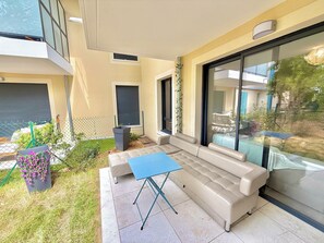 Ein moderner Sitzbereich im Freien mit einer Couch und einem Tisch, eine Glastür, die auf einen Balkon führt, und ein kleiner Garten mit Blumen.