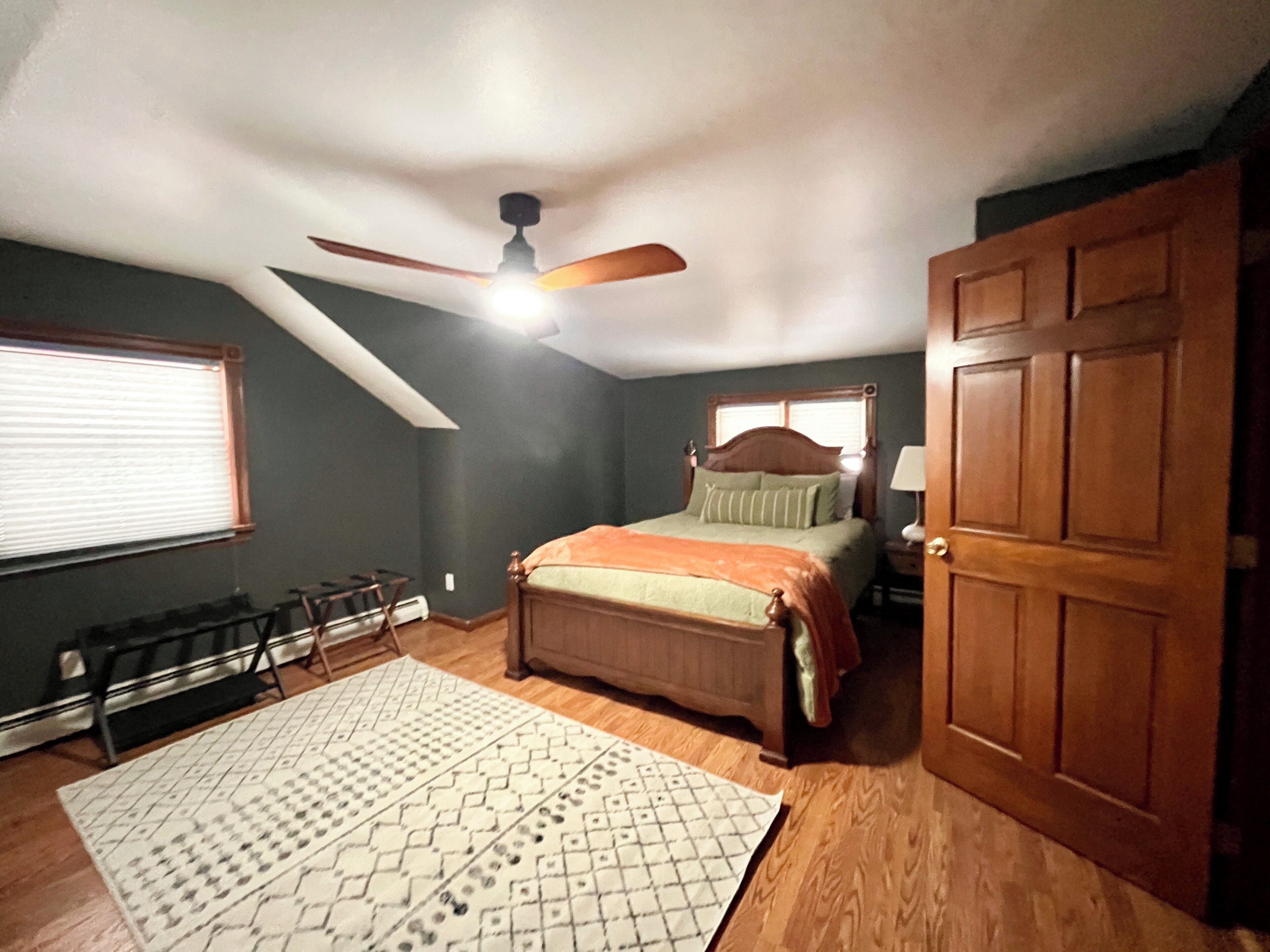 Une chambre à coucher avec un lit, un ventilateur de plafond, une fenêtre avec des stores, une porte et un tapis à motifs.