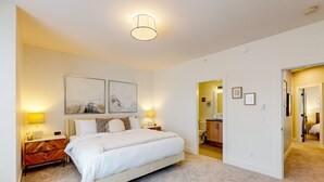Un dormitorio con cama, lámparas de noche, mesita de noche, un cuadro colgado en la pared y una puerta que da a otra habitación.