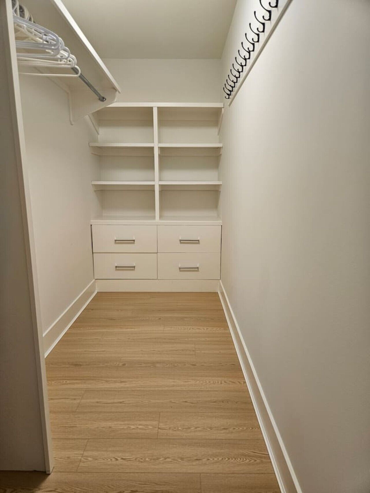 Um closet estreito com espaço para passar, piso de madeira, paredes brancas e uma estante embutida com vários compartimentos.