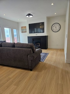 A living room with a sofa, a fireplace, a flat-screen TV, and a wall clock. - Luxe 4BD family retreat w/AC, Yard, Bunks and King (Calgary)