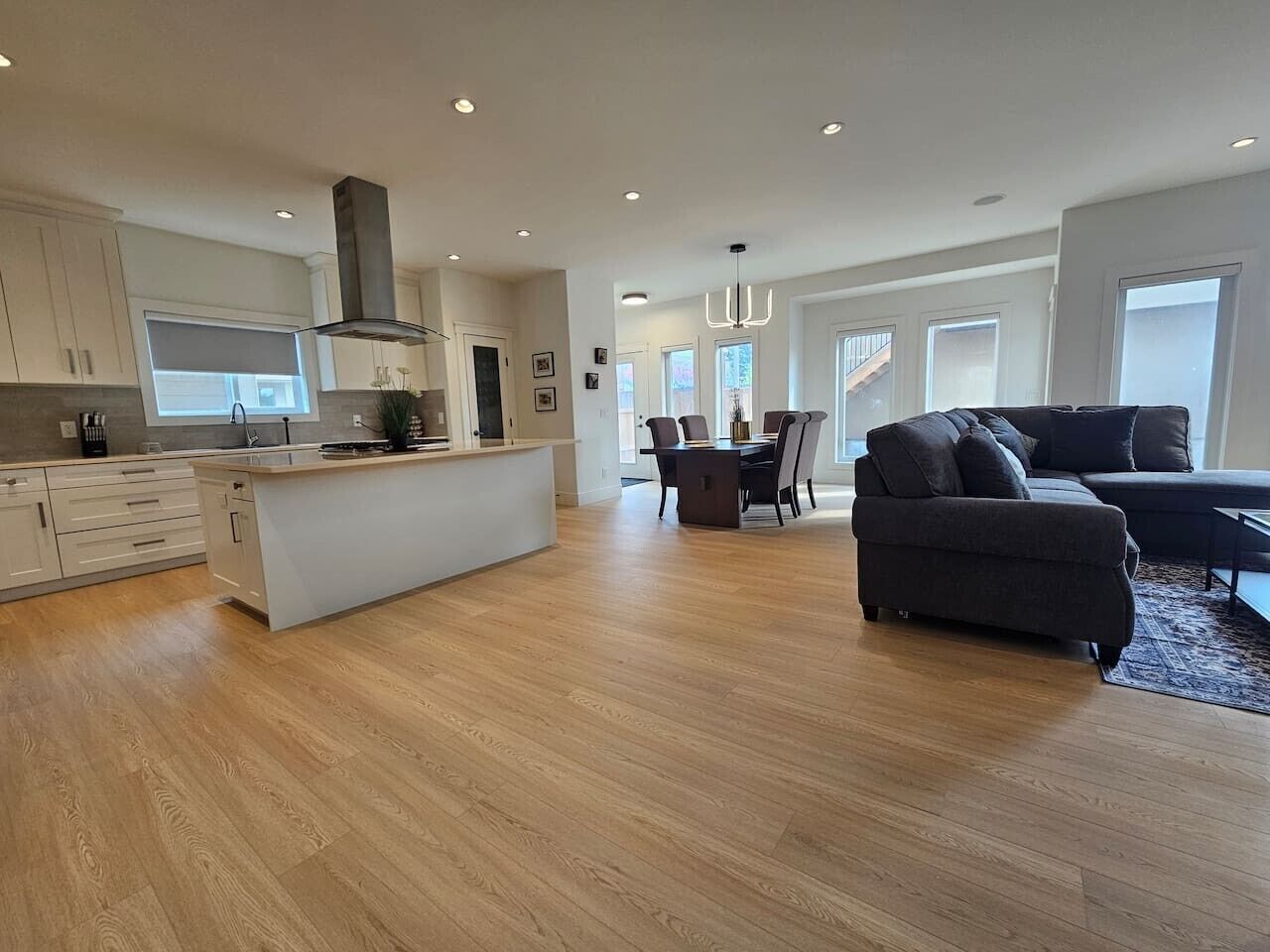 A spacious living area with a dark-colored sofa and a dining table, a kitchen with white cabinets and a central island, and a large window providing natural light.