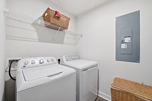 A laundry room with two white front-loading washers and dryers, a metal shelf with baskets, and a grey electrical panel. - Modern 3BR Townhome- Perfect for Short/Long Stays- Vacation or Traveling Pros! (Myrtle Beach)