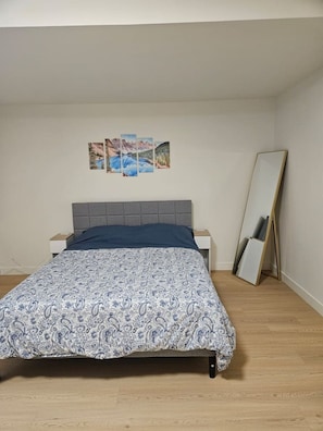 Ein Schlafzimmer mit einem Bett, einem Nachttisch, einem Wandbild und einem stehenden Spiegel.