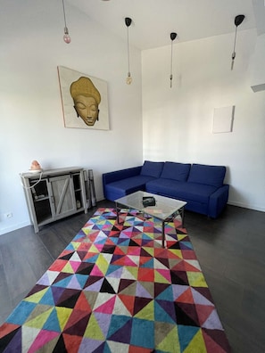 A living room with a blue sofa, a glass coffee table, a colorful geometric rug, a cabinet, and a Buddha statue. - Bright apartment with 2 balconies – family-friendly – Beach and shops within walking distance (SEIGNOSSE)