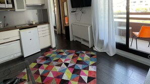 Eine moderne Küche mit weißen Schränken, einem eingebauten Ofen und einer Mikrowelle. Auf dem Boden liegt ein farbenfroher geometrischer Teppich, an der Wand ist ein Flachbildfernseher angebracht und es gibt ein Fenster mit weißem Vorhang.