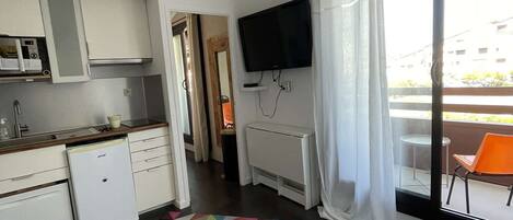 A modern kitchen with white cabinets, a built-in oven, and a microwave. There's a colorful geometric rug on the floor, a flat-screen TV mounted on the wall, and a window with a white curtain.