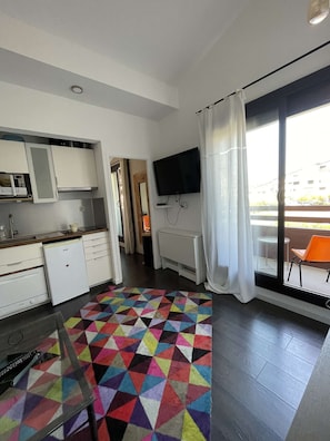 A modern kitchen with white cabinets, a built-in oven, and a microwave. There's a colorful geometric rug on the floor, a flat-screen TV mounted on the wall, and a window with a white curtain. - Bright apartment with 2 balconies – family-friendly – Beach and shops within walking distance (SEIGNOSSE)