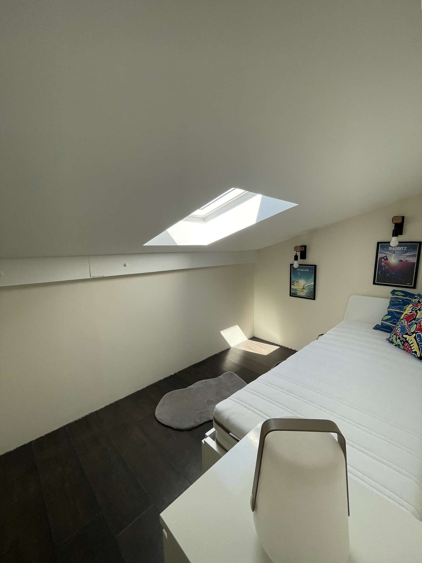 A bedroom with a skylight, a bed, a pillow, a small rug, and a wall-mounted light fixture.
