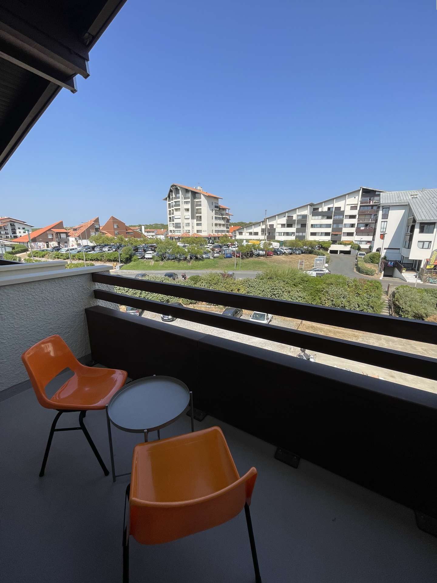 A balcony with two orange chairs and a small table, overlooking a parking lot and buildings.