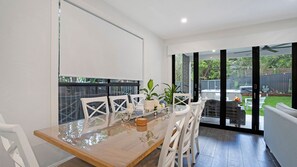 A dining area with a glass table, white chairs, and a view of a backyard with a barbecue and outdoor furniture. - Salt & Spa Norah Head (Norah Head)