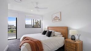 A bedroom with a bed, a nightstand, a lamp, a ceiling fan, a window with a view, and a framed picture on the wall. - Salt & Spa Norah Head (Norah Head)