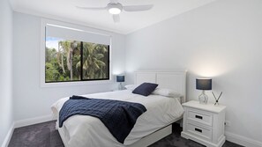 A bedroom with a bed, a nightstand, a lamp, a ceiling fan, and a window with a view of trees. - Salt & Spa Norah Head (Norah Head)