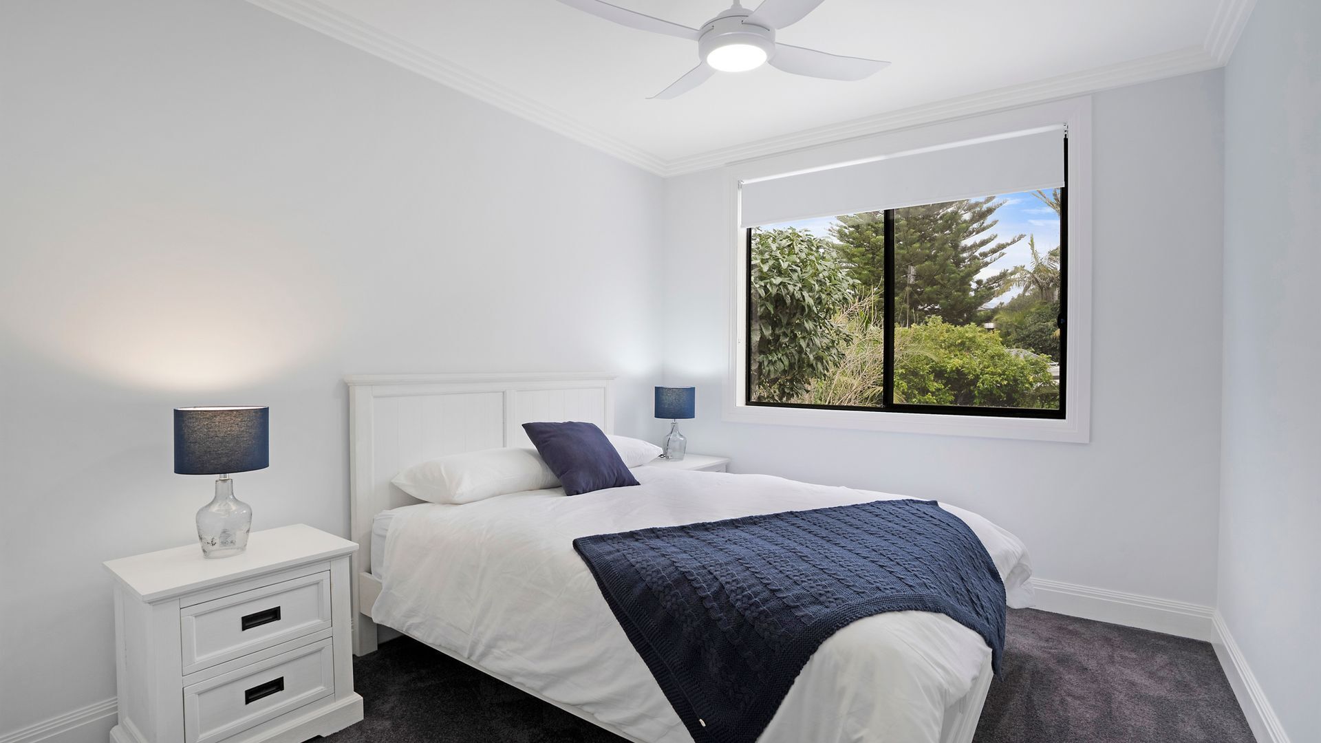 Une chambre à coucher comprenant un lit, des tables de chevet, un ventilateur de plafond, une fenêtre donnant sur un espace verdoyant et une moquette sombre.