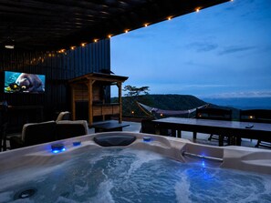 Un jacuzzi con vistas a un paisaje montañoso, una estructura de madera con un televisor y mobiliario exterior.